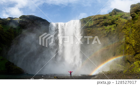 Unidentified woman stands beneath the powerful Skogafoss waterfall in Iceland, framed by mist and a vivid rainbow Unidentified woman stands beneath the powerful Skogafoss waterfall in Iceland, framed by mist and a vivid rainbow 128527017