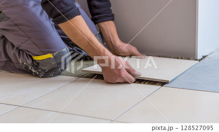 A master who is laying ceramic tiles on the floor during a renovation or construction project. The master is on his knees, carefully aligning the tiles and applying the adhesive mixture.	 128527095