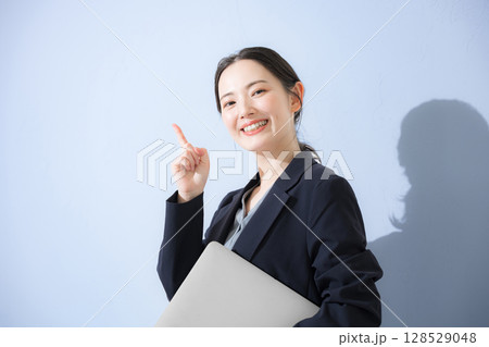 パソコン持ってスーツを着た立つポイントなどの指差しポーズの若いかわいい女性のビジネス上半身　水色背景 128529048