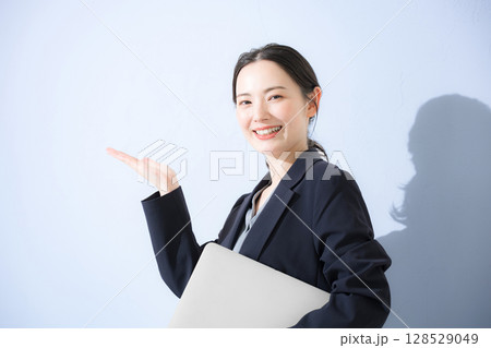 パソコン持ってスーツを着た立つオススメや案内のポーズ若いかわいい女性のビジネス上半身 水色背景の営業 パソコン持ってスーツを着た立つオススメや案内のポーズ若いかわいい女性のビジネス上半身 水色背景の営業 128529049