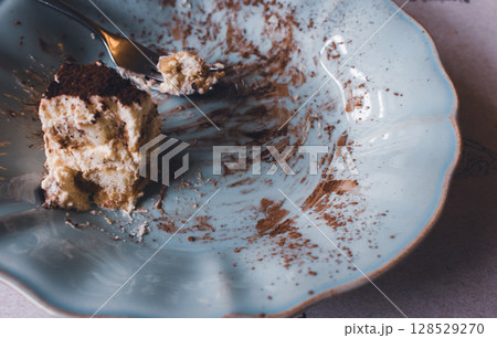 An uneaten piece of cake on blue vintage plate. Slice of tiramisu cake with fork on plate, close up. Delicious chocolate biscuit for breakfast. Sweet food concept. Italian dessert. An uneaten piece of cake on blue vintage plate. Slice of tiramisu cake with fork on plate, close up. Delicious chocolate biscuit for breakfast. Sweet food concept. Italian dessert. 128529270