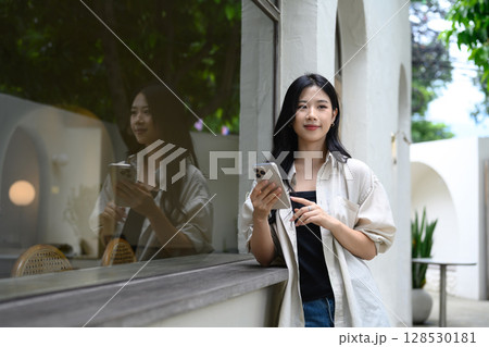 Stylish young woman standing outside a cafe while holding her smartphone 128530181