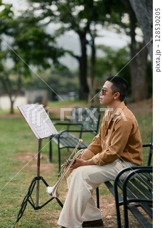 Trumpet player resting on bench with music stand, enjoying a quiet outdoor practice session Trumpet player resting on bench with music stand, enjoying a quiet outdoor practice session 128530205
