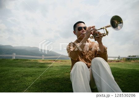 Man in sunglasses playing trumpet sitting on green grass with scenic mountain backdrop Man in sunglasses playing trumpet sitting on green grass with scenic mountain backdrop 128530206