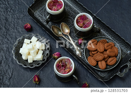 Floral tea made from rose petals and cookies on a dark gray concrete background. Tea drinking in eastern style. Romantic breakfast for Valentine's Day, Women's Day, Mother's Day Floral tea made from rose petals and cookies on a dark gray concrete background. Tea drinking in eastern style. Romantic breakfast for Valentine's Day, Women's Day, Mother's Day 128531703