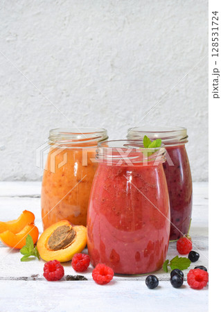 Orange, red and purple smoothies of fresh ingredients - bananas, apricots, raspberries and blueberries on a light background. Healthy food concept. Vegetarian product. Organic Raw Drink. Copy space. 128531724