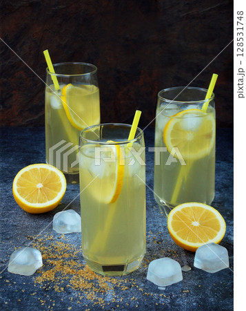 Three glasses of cold homemade lemonade with lemon slices, ice cubes, brown sugar and straws on dark background. Copy space Three glasses of cold homemade lemonade with lemon slices, ice cubes, brown sugar and straws on dark background. Copy space 128531748