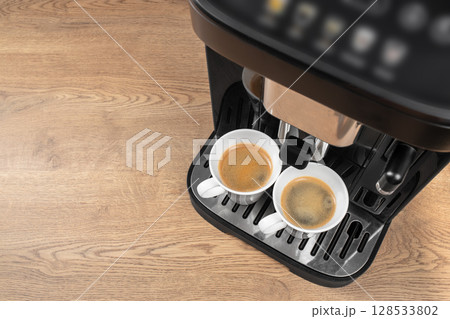 A modern coffee machine brews two fresh espressos into white cups on a wooden countertop from a top-down perspective A modern coffee machine brews two fresh espressos into white cups on a wooden countertop from a top-down perspective 128533802