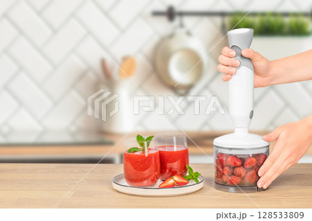 Hands operate a hand blender filled with strawberries next to a plate with two glasses of fresh strawberry smoothie garnished with mint on a wooden kitchen counter 128533809
