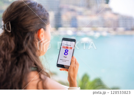 Young woman checking UV index on smartphone with high radiation alert. Mobile app warns to apply sunscreen before going outdoors 128533823