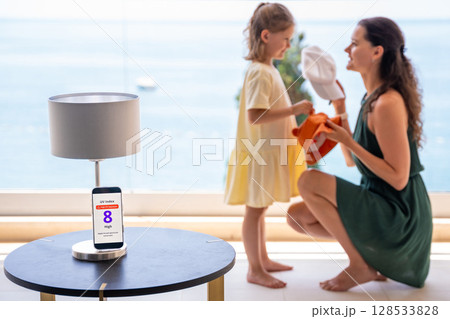 Smartphone shows high UV alert while mother offers child a cap or sun hat. Everyday sun protection choices in response to radiation warning 128533828