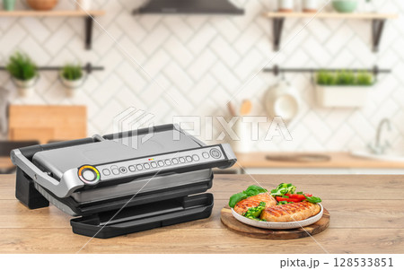 A modern electric grill sits on a wooden countertop beside a plate of freshly grilled chicken breasts with herbs and vegetables in a cozy kitchen 128533851