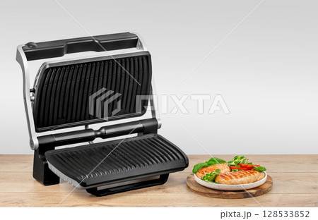 An open electric grill is displayed next to a serving plate of freshly grilled chicken breasts with vegetables on a wooden kitchen surface. copy space An open electric grill is displayed next to a serving plate of freshly grilled chicken breasts with vegetables on a wooden kitchen surface. copy space 128533852