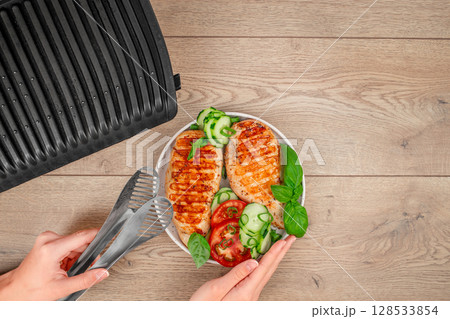 A top-down view shows hands preparing grilled chicken with cucumbers, tomatoes, and basil next to an open electric grill on a wooden surface. copy space A top-down view shows hands preparing grilled chicken with cucumbers, tomatoes, and basil next to an open electric grill on a wooden surface. copy space 128533854