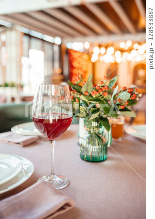 Elegant restaurant table setting with a glass of red wine and a floral centerpiece in warm ambient lighting Elegant restaurant table setting with a glass of red wine and a floral centerpiece in warm ambient lighting 128533887
