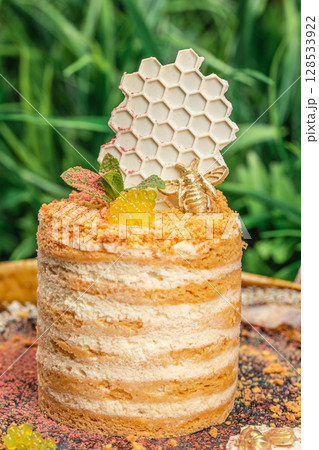 Elegant honey cake with layered cream and sponge, topped with white chocolate honeycomb, golden bee, mint leaves, and decorative spheres, set against a natural green background 128533922