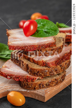 Stacked rye bread slices topped with spicy pork belly, fresh spinach leaves, and cherry tomatoes, served on a rustic wooden board Stacked rye bread slices topped with spicy pork belly, fresh spinach leaves, and cherry tomatoes, served on a rustic wooden board 128533924