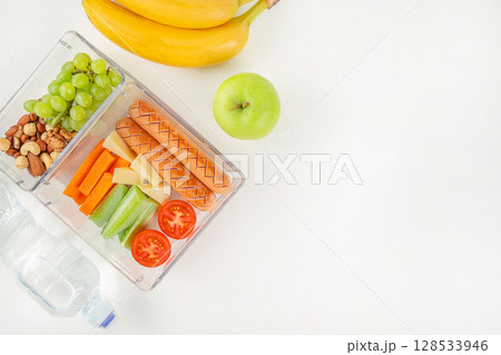 Balanced lunchbox with sausages, cheese, vegetables, grapes, nuts, and water, arranged with fresh fruit on a bright white background. top view. copy space. 128533946