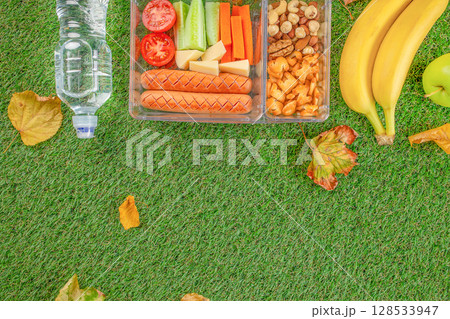 Healthy picnic-style lunch with sausages, vegetables, cheese, nuts, crackers, and fruit in a container on green grass with autumn leaves and a bottle of water 128533947
