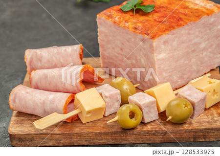 Rolled ham slices, cheese cubes, olives, and skewered appetizers are arranged on a wooden board next to a large block of ham loaf for a hearty and inviting snack display 128533975
