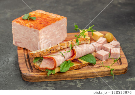 A hearty sandwich with rolled ham slices, cheese, and greens is served on a wooden board next to a block of ham loaf and diced ham cubes for a rustic and satisfying meal A hearty sandwich with rolled ham slices, cheese, and greens is served on a wooden board next to a block of ham loaf and diced ham cubes for a rustic and satisfying meal 128533976