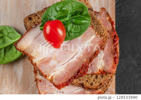 Close-up of an open sandwich with spicy pork belly on rye bread, garnished with a fresh spinach leaf and cherry tomato 128533990