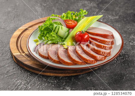 Sliced smoked sausage is elegantly arranged on a plate with fresh lettuce, cucumber ribbons, tomato, and red onion for a vibrant and appetizing presentation 128534070