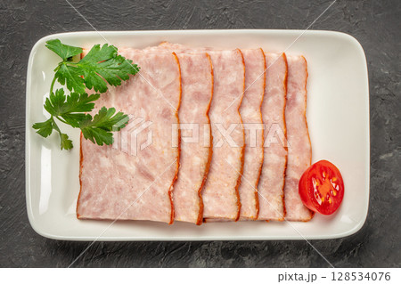 Thin slices of cooked ham are neatly arranged on a white rectangular plate, garnished with fresh parsley and a halved cherry tomato for a clean, appetizing presentation Thin slices of cooked ham are neatly arranged on a white rectangular plate, garnished with fresh parsley and a halved cherry tomato for a clean, appetizing presentation 128534076