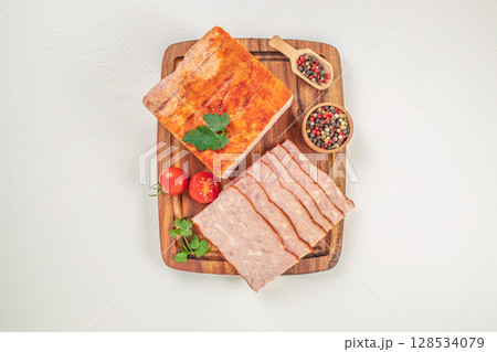 Sliced ham loaf is displayed on a wooden board with whole and halved cherry tomatoes, parsley, and colorful peppercorns, creating a simple and appetizing presentation 128534079