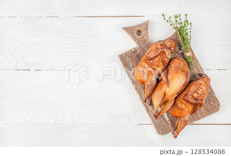 Three whole smoked quails are beautifully arranged on a rustic wooden cutting board with fresh thyme, creating a simple yet elegant presentation of gourmet poultry 128534086