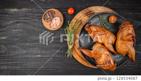Three perfectly smoked quails are artfully arranged on a dark rustic plate with fresh thyme, coarse salt, and colorful peppercorns, creating a rich and inviting gourmet presentation 128534087