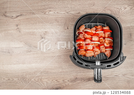 Raw chicken and vegetable skewers are neatly arranged inside an air fryer basket, ready for healthy and convenient cooking on a wooden kitchen countertop 128534088