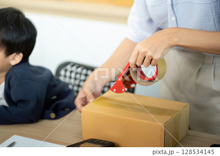 Sell Online: A mother sealing packages for her e-commerce business with her child nearby Sell Online: A mother sealing packages for her e-commerce business with her child nearby 128534145