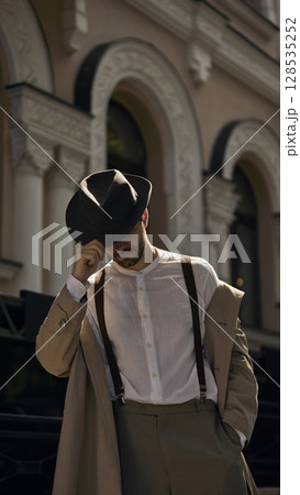 Man in vintage attire tips hat in front of ornate building with arched openings Man in vintage attire tips hat in front of ornate building with arched openings 128535252