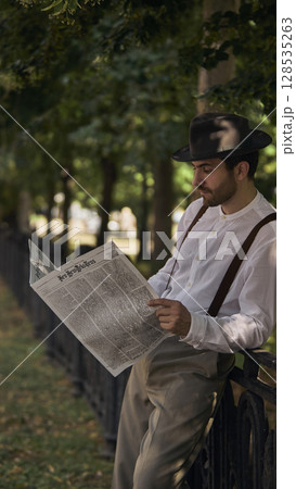 Man reading newspaper in vintage attire leaning against park fence under trees 128535263