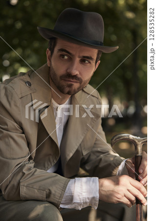 Man in vintage attire sitting on park bench under trees holding cane Man in vintage attire sitting on park bench under trees holding cane 128535282