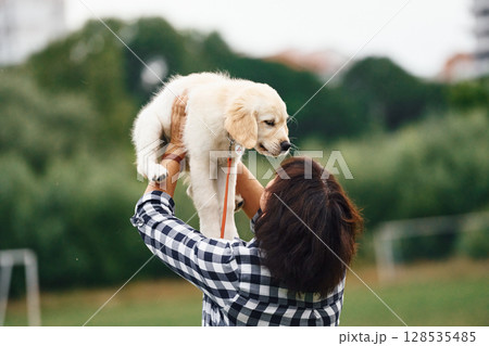 Puppy is in hands. Woman with cute little golden retriever dog is on the green field 128535485