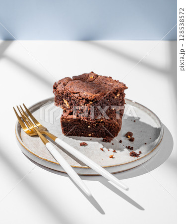 Chocolate brownie with nuts cut into pieces on a plate on light background with cutlery and shadows. Chocolate brownie with nuts cut into pieces on a plate on light background with cutlery and shadows. 128538572