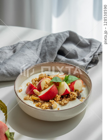Natural greek yogurt with granola and peach in a bowl on a white table near window 128538590