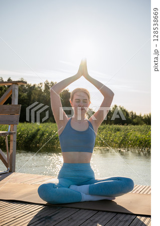 Young woman practicing yoga pose on wooden dock near lake in summer morning light 128538669