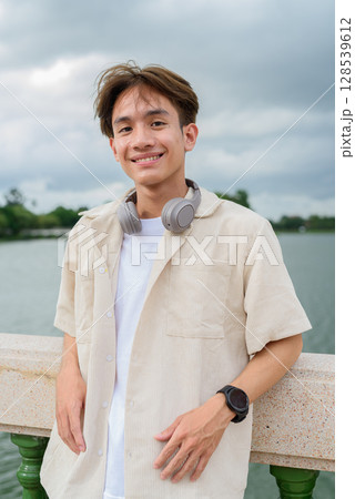 Asian non binary tourist person with headphones relaxing at park near lake 128539612