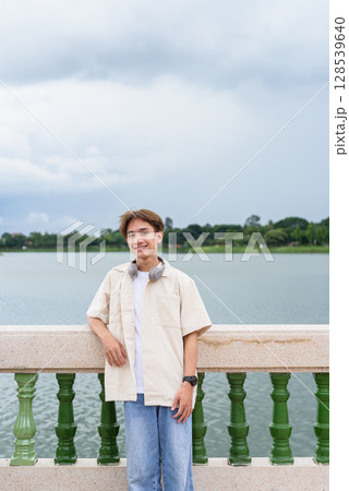 Asian non binary tourist person with headphones relaxing at park near lake Asian non binary tourist person with headphones relaxing at park near lake 128539640