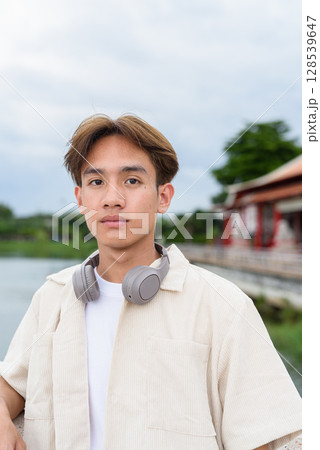 Asian non binary tourist person with headphones relaxing at park near lake 128539647