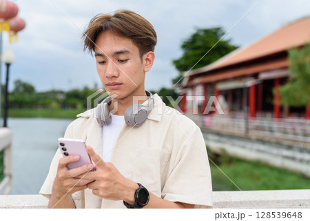 Asian non binary tourist person with headphones relaxing at park near lake using phone 128539648