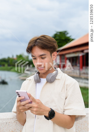 Asian non binary tourist person with headphones relaxing at park near lake using phone 128539650
