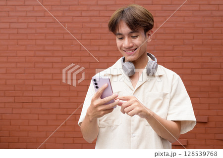 Non binary cute Asian young gay man using mobile phone against brick wall 128539768