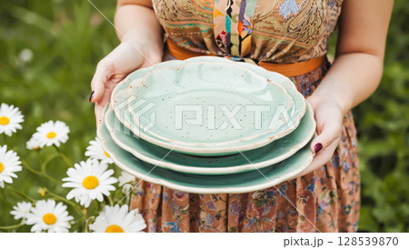 Woman Holding Stack Of Green Ceramic Plates Outdoors Near Chamomile Flowers For Tableware Visuals. Concept Of Handmade Pottery, Outdoor Gatherings, Country Kitchen Style, And Summer Entertaining 128539870