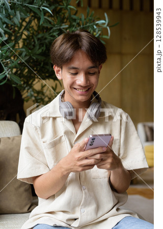 Non binary Asian student man sitting in modern coffee shop texting with phone 128539943