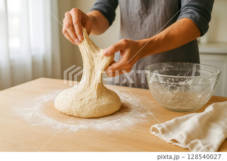 Stretching Homemade Bread Dough On Wooden Table With Flour For Artisan Baking And Slow Food Visuals. Concept Of Natural Fermentation, Handcrafted Bread Making, Home Cooking, And Traditional Recipes Stretching Homemade Bread Dough On Wooden Table With Flour For Artisan Baking And Slow Food Visuals. Concept Of Natural Fermentation, Handcrafted Bread Making, Home Cooking, And Traditional Recipes 128540027