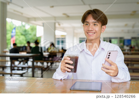 Non binary university student sitting in college campus using phone Non binary university student sitting in college campus using phone 128540266